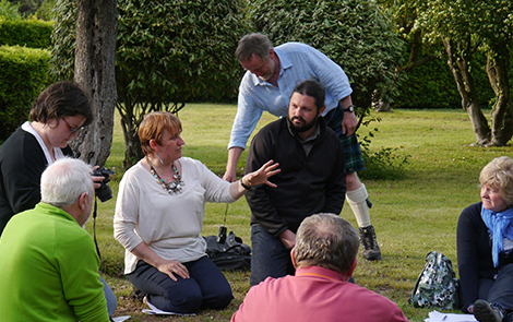 A group of 6 people sat on the lawn at the Italian President's summer residence, participating in a workshop for Interpret Europe, being led by Susan Cross. One man in a kilt is stood in the background putting a camera on the ground.
